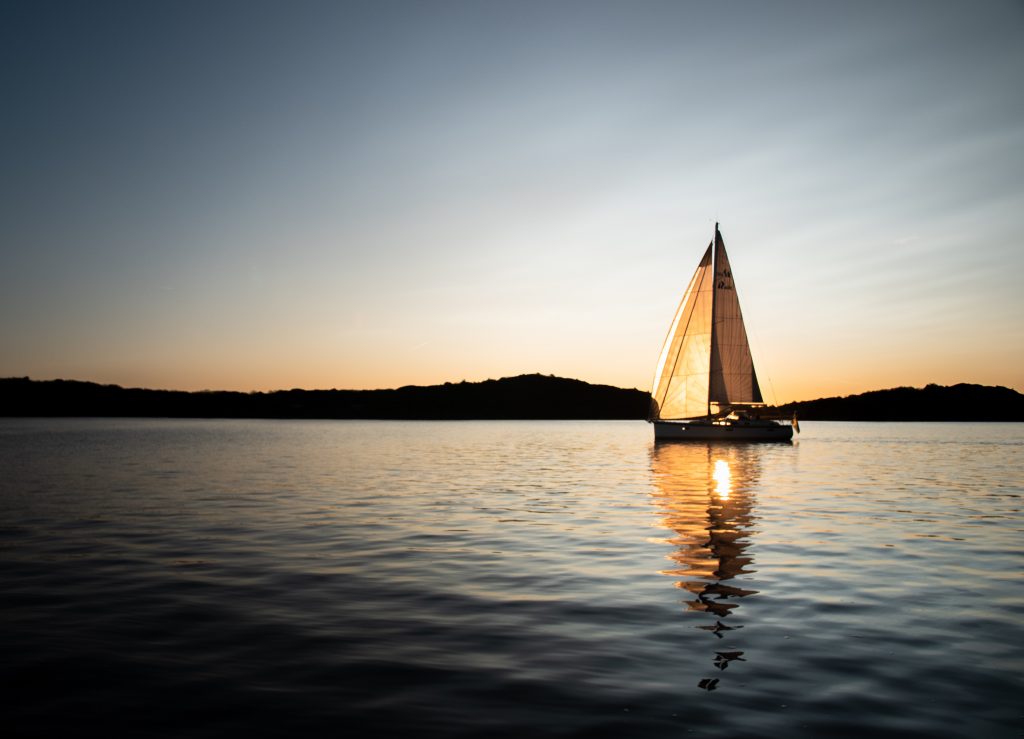 8 tipi di amici da non portare con sé durante una vacanza in barca al tramonto in mare aperto, in lontananza si vedono delle isole e l'acqua del mare è molto piatta e calma